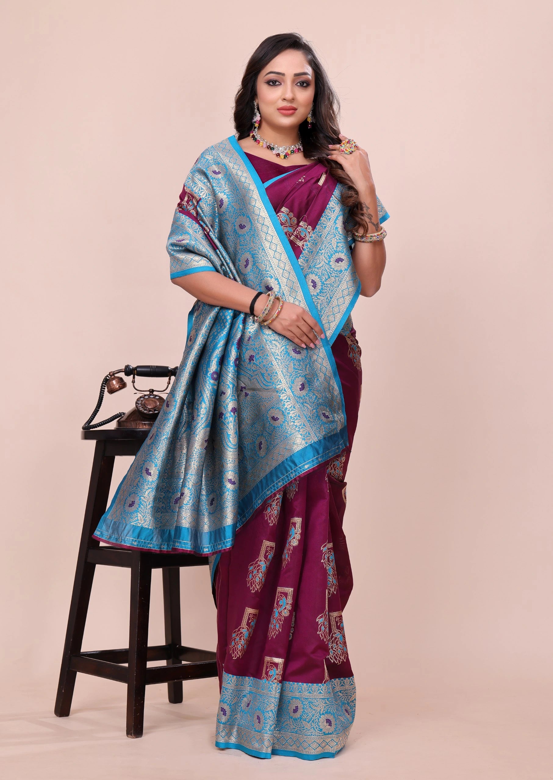 Woman wearing a purple saree with blue border, sitting on a stool against a beige background
